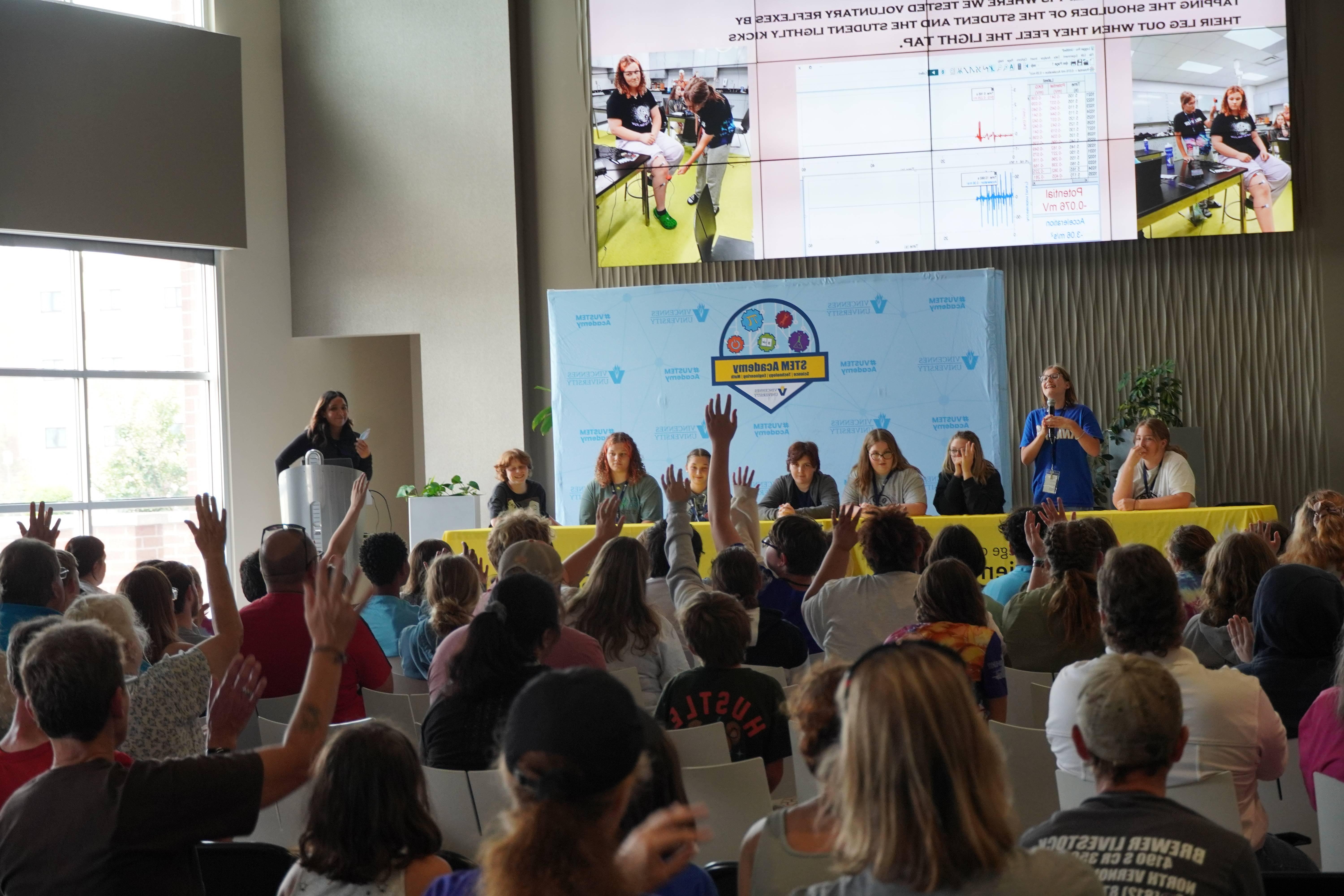 Male and female STEM Academy students sitting and standing at a long table on a stage. Other students sit in chairs in front of the stage.